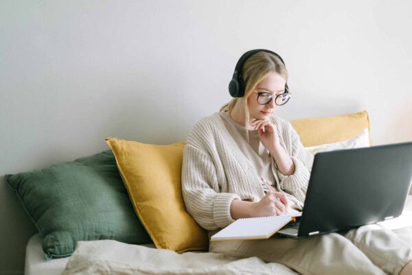 A woman sitting on a couch/bed while working on a laptop and writing on a notebook.